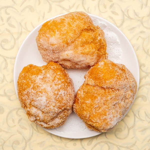 Three golden-brown sugar-dusted donuts on a white plate with a decorative doily, against a patterned yellow background.