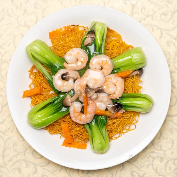 Crispy egg noodles topped with shrimp, bok choy, mushrooms, and carrots on a white plate.